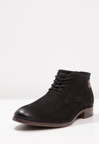 Bottine en daim noir avec bout arrondi, lacets à l'avant et détails discrets de couture. Talon en bois qui contraste avec l'ensemble du design.