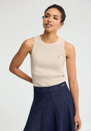 Vrouw met kort krullend haar, die een beige geribde mouwloze top en een donkerblauwe hoge taille rok draagt, staand tegen een effen achtergrond.