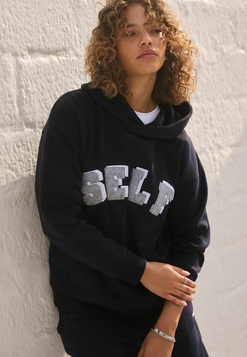 Schwarzer Kapuzenpullover mit großem, strukturiertem "SELF"-Schriftzug in Grau, aus weichem Baumwollstoff. Modell lehnt an einer weißen Wand.