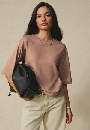 Młoda kobieta z długimi ciemnymi włosami, ubrana w luźny brązowy t-shirt, beżowe spodnie, złoty łańcuszek, bransoletkę i trzymająca czarną skórzaną torbę.