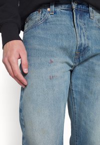 Jeans de mezclilla azul claro con un diseño clásico, estilo de cinco bolsillos, y signos visibles de uso con manchas y desvanecimiento en la tela.