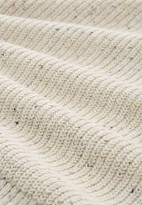 Cremefarbener gestrickter Stoff mit gerippter Textur, der kleine schwarze und braune Flecken aufweist, in Nahaufnahme mit Wellen und Falten dargestellt.