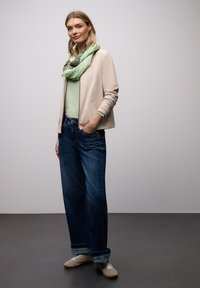 Femme debout les mains dans les poches portant une veste beige clair, une écharpe verte à motifs, un haut vert, un jean bleu à jambes larges et des baskets beige.