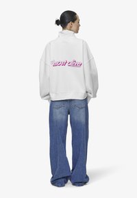 Sudadera blanca de gran tamaño con cuello alto, con la frase "más vivo" en letras rosas en la parte posterior. Combinada con jeans azules de corte holgado.
