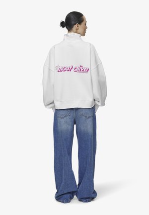 Felpa bianca oversize con colletto alto, con la scritta "most alive" in lettering rosa sul retro. Abbinata a jeans blu dal fit morbido.