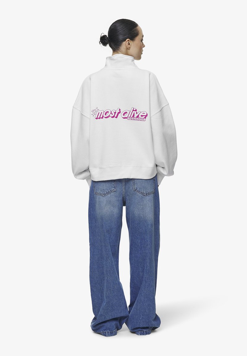 Sudadera blanca de gran tamaño con cuello alto, con la frase "más vivo" en letras rosas en la parte posterior. Combinada con jeans azules de corte holgado.