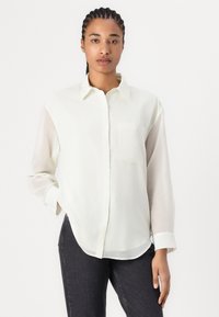 Knappe skjorte i lysecreme stof, med lange gennemsigtige ærmer, en brystlomme og en buet kant, parret med mørke jeans.