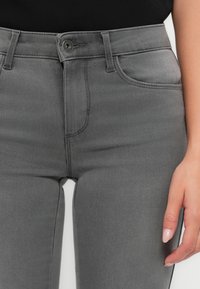 Jeans de mezclilla gris con un ligero estiramiento. Presenta bolsillos frontales, un cierre de botón y una textura suave. Acentuado con costuras limpias.
