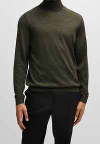 Mężczyzna w ciemnooliwkowym golfie i czarnych spodniach, stojący na tle jasnym.