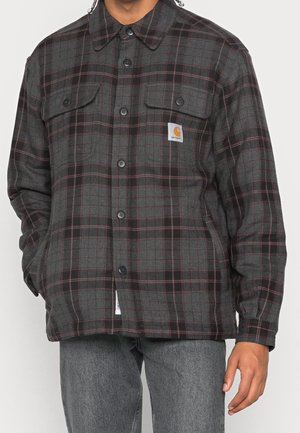 Homme portant une chemise à carreaux gris foncé et noir à boutons avec poches poitrine et écusson logo Carhartt, assortie à un jean gris.
