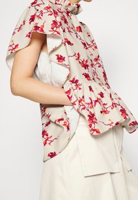 Blusa floreale color crema con motivi rosa e rossi, maniche arricciate, colletto alto e vita a peplo, abbinata a una gonna di colore chiaro.