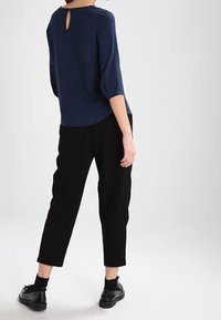 Blusa blu navy con dettaglio a occhio di bue sul retro, abbinata a pantaloni neri morbidi e scarpe nere, creando una silhouette snella.