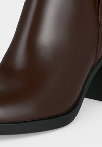 Bottine en cuir lisse marron foncé avec bout arrondi et talon bloc bas noir sur fond blanc.
