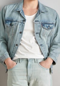 Ljusblå denimjacka med två bröstfickor och mörka knappar, i kombination med en vit T-shirt och matchande ljusblå jeans.