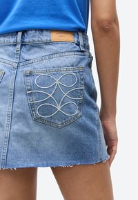 Svijetloplava traper mini suknja s bijelim izvezenim petljama na stražnjem džepu, nošena s uvučenom plavom košuljom, prikazuje donji dio trupa i ruku.