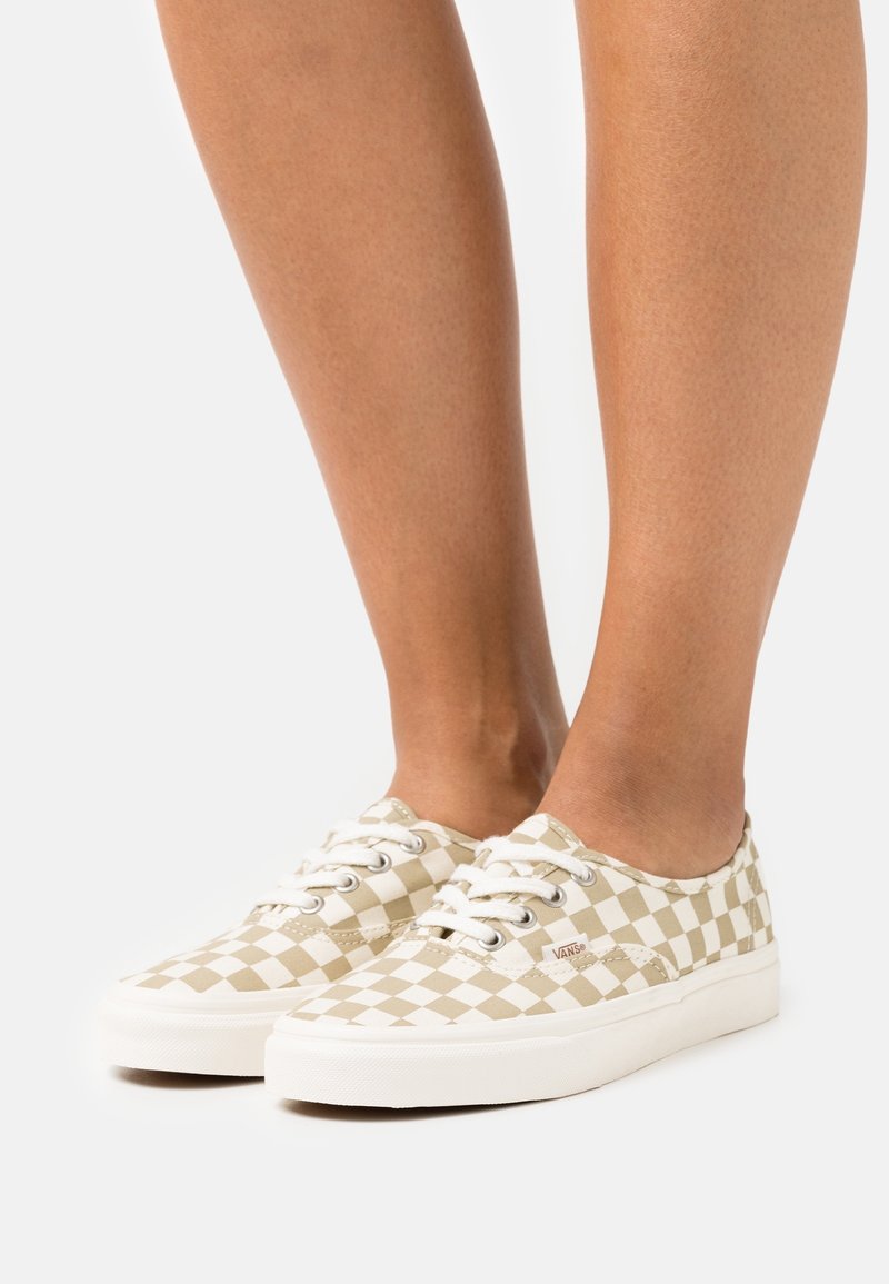 Ben som bär låga beige och vita rutiga Vans sneakers med vita snören och gummisulor mot en vit bakgrund.