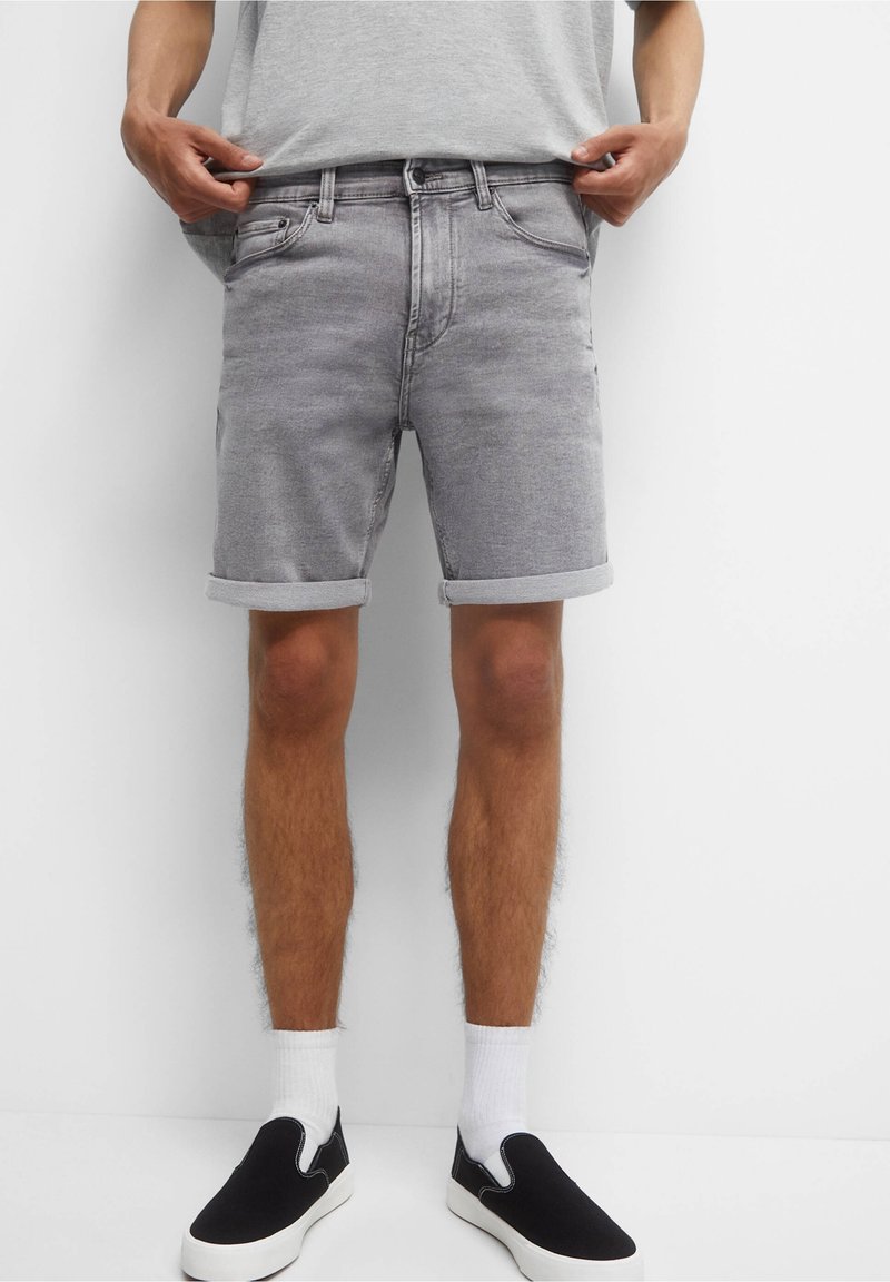 Grijze denim shorts met omgeslagen cuffs, vijf zakken en een rits. Gemaakt met zwarte instappers en witte sokken. Eenvoudig grijs t-shirt erboven.