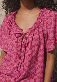 Lyserød bluse med mønster af blomster og krabber, med en rynket halsudskæring, bindedetalje og frynser. Blød tekstur, afslappet pasform, korte ærmer.