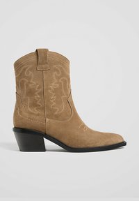 Botte de cowboy en suede marron clair avec des motifs de couture courbes, un bout pointu et un talon empilé noir. Comprend une languette pour un enfilage facile.