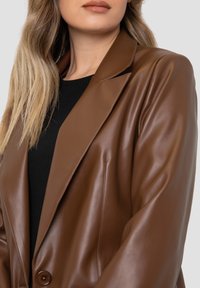 Brown faux leather blazer with a notched lapel, single button closure, and tailored fit. Smooth texture and structured design details.