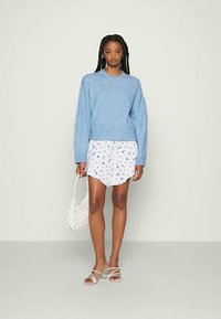 Mulher de pé, vestindo um suéter azul claro, uma saia mini branca com flores, sapatos de salto brancos com tiras, e segurando uma bolsa de palha branca.