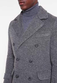 Abrigo de lana gris de mezcla con doble botonadura y acabado texturizado, cuello de muesca, botones negros y un bolsillo en el pecho. Estilizado sobre un cuello de tortuga gris.