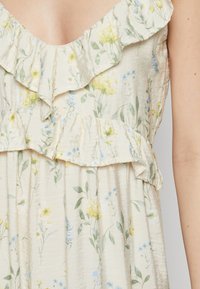 Robe fleurie avec un fond crème, ornée de fleurs jaunes et bleues. Encolure volantée et volants en tissu léger et texturé.