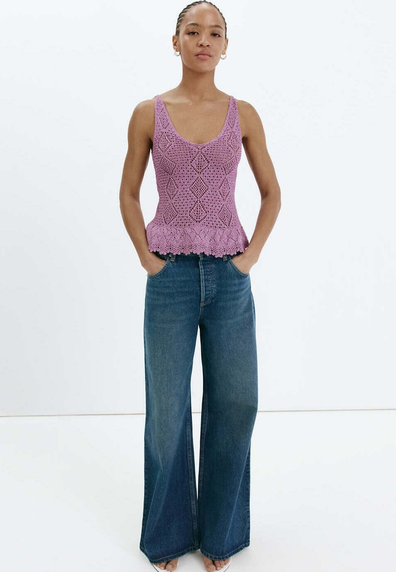Femme portant un haut sans manches violet en crochet et un jean bleu à jambes larges, debout les mains dans les poches devant un fond uni.