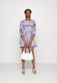 Vestido azul com padrão, mangas três quartos e bainha flared. Bolsa tecida branca segurada nas mãos, sapatos de plataforma cinzentos nos pés.