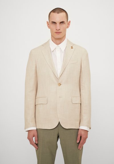 Blazer di lino beige con un solo bottone, due tasche frontali e una spilla decorativa sul colletto. Indossato sopra una camicia bianca e pantaloni verdi.