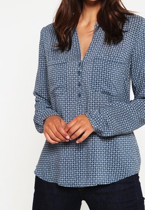 Nainen, joka pukeutunut pitkähihainen sininen paita, jossa valkoinen ympyräkuvio, kaksi rintataskua ja tummat housut, seisoo kädet ristissä.