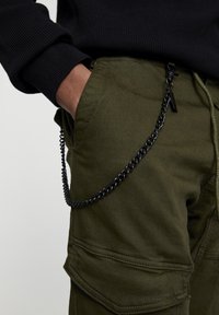Groene cargobroek met een gestructureerde afwerking, voorzien van een zwart kettingaccessoire bevestigd aan de zak en een geribde zwarte trui-mouwomslag.