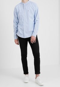 Light blue button-up shirt with a stand collar, chest pocket; paired with black pants and white sneakers, presented in a neutral background.