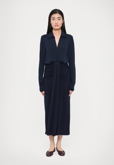 Navy long-sleeve dress with a collared neckline, ruched waist detail, and a straight skirt. Model wears dark flat shoes. Smooth fabric texture.