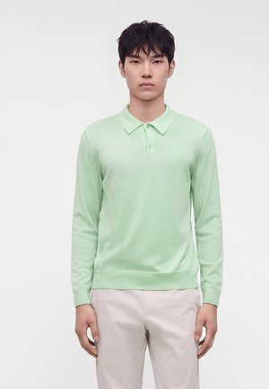 Jeune homme aux cheveux noirs courts portant un polo vert clair à manches longues et un pantalon beige sur un fond blanc uni.