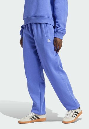 Osoba ubrana w niebieskie dresy Adidas i pasującą bluzę, stojąca w białych tenisówkach Adidas z czarnymi paskami i gumowymi podeszwami.