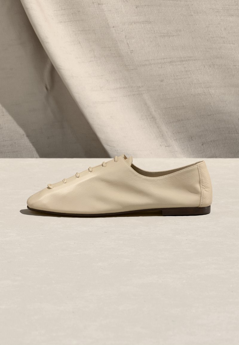 Chaussure plate en cuir beige avec bout arrondi, lacets de style bouton et semelle foncée sur une surface claire texturée avec une ombre douce en arrière-plan.