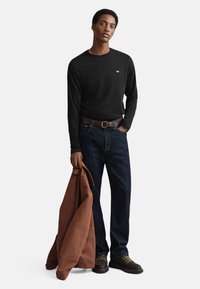 Black long-sleeve shirt, dark blue jeans, brown leather belt, holding a brown jacket. Black shoes with laces. Simple design, no patterns.