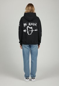 Felpa nera con scritta bianca "MI AMOR" e grafica sul retro, abbinata a pantaloni in denim azzurro chiaro e sneakers nere.