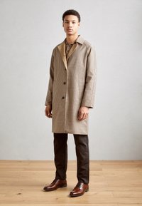 Manteau à carreaux marron avec un col cranté, associé à une chemise beige et un pantalon foncé, complété par des chaussures en cuir marron. Fond en parquet bois.
