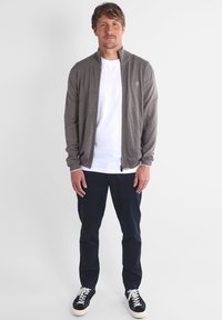 Homme portant une veste zippée grise sur un t-shirt blanc, un pantalon foncé et des baskets noires, adossé à un fond clair et uni.