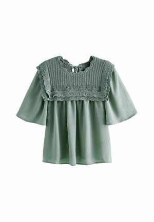 Kobieta w jasnooliwkowej bluzce z szerokim, dzianym kołnierzem oraz długiej, niebieskiej dżinsowej spódnicy z rozcięciem z przodu i białych, frędzlowanych botkach za kostkę.