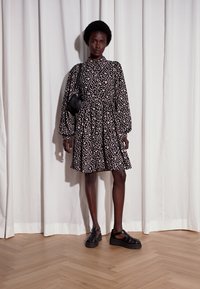 Vestido preto com padrão floral, colarinho, mangas volumosas longas e saia rodada. Usado com sandálias pretas de tiras e uma pequena bolsa preta.