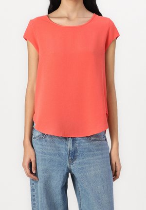 Kvinde iført en koral kortærmet bluse stoppet ned i lyseblå højtaljede jeans, stående mod en ensfarvet hvid baggrund.