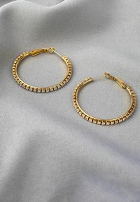 Gold hoop earrings with a textured edge, featuring a row of small clear crystals along the bottom half. Set against a light gray fabric background.