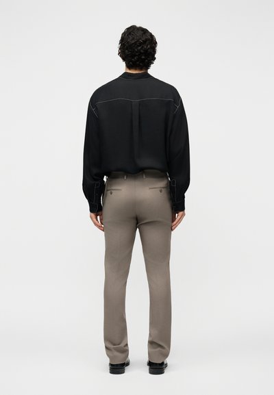 Homme aux cheveux bouclés et foncés, de dos, portant une chemise noire à manches longues, un pantalon beige et des chaussures noires, sur un fond uni.