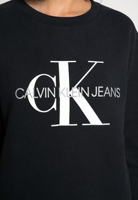 Sudadera negra con un gran logo blanco "CK" y el texto "CALVIN KLEIN JEANS" debajo. Material de algodón suave con diseño de cuello redondo.