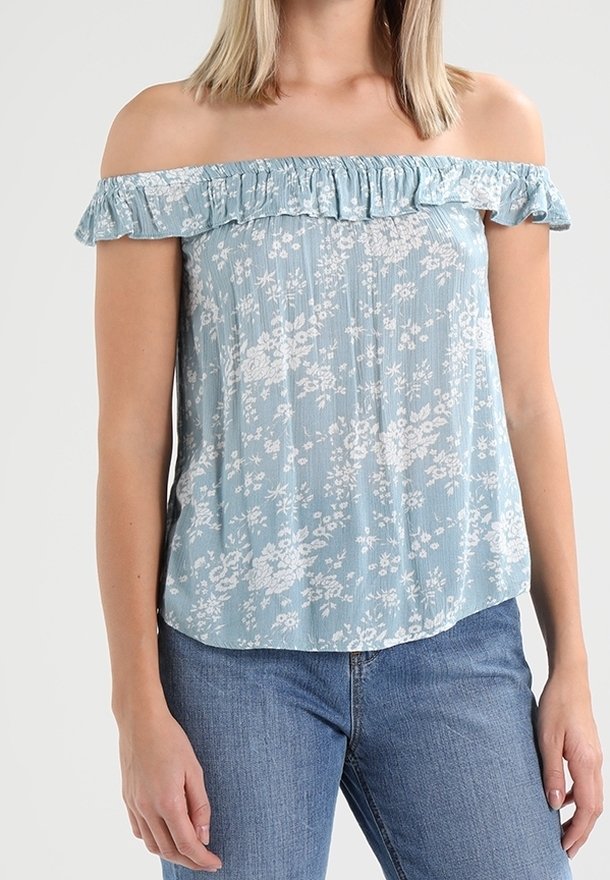 Kobieta w jasnoniebieskim topie odkrywającym ramiona z białym kwiatowym wzorem i niebieskich dżinsach, stojąca na neutralnym tle.