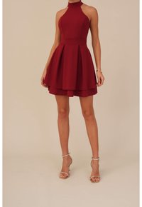 Punainen hihaton mekko, jossa on korkea kaulus, tiukka vartalo ja levenevä hame. Valmistettu sileästä kankaasta, jossa on kerroksellinen helma. Hopeiset nilkkaremmit.