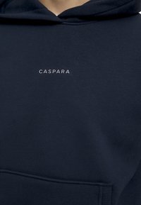 Navy blå hettegensere laget av mykt stoff, med en fremre lomme og en liten, lys grå logo som leser "CASPARA" på brystet.